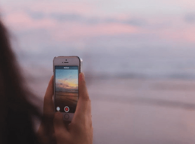 Henkilö kuvaa auringonlaskua rannalla älypuhelimella. 