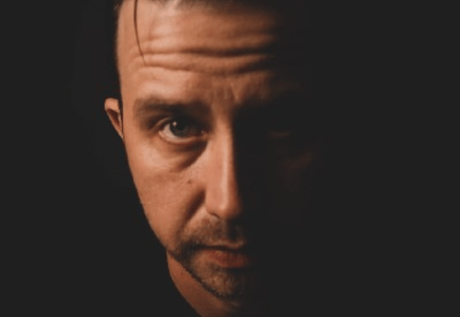 Markus Råhman katsoo kameraan tummaa taustaa vasten. 