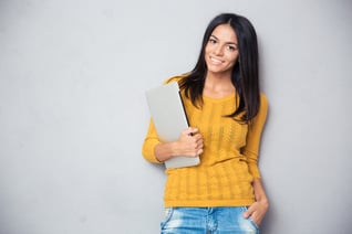 Portrait of a happy casual woman holding laptop over gray background and looking at camera